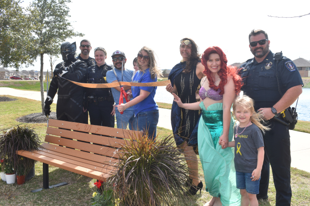 Amanda Torres, center, is surrounded by family and superhero friends as she cuts a ribbon to a bench dedicated to her late son, DJ Pruitt. Photo by Katherine Anthony