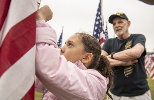 8-year-old Lourdes Aviña places a name tag on a flag under the watchful eye of Vietnam veteran Mike Wright  at the Field of Honor on Saturday, November 4, 2023.