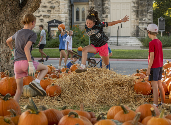 Margaret Bruechner, facing camera, and Adeline Lake, left, both 10, enjoyed playing in aromatic hay while helping unload pumpkins at First United Methodist Church on Wednesday, October 9, 2024.  2,880 large pumpkins, plus many smaller versions were unloaded at the church.   The pumpkins were grown in New Mexico.   A second, but smaller batch of pumpkins will arrive from there on October 19th.   Sales continue through 5p.m. on October 31st.    Photo by Andy Sharp.
