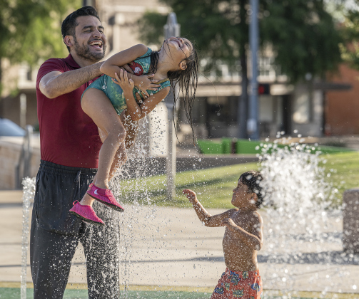 Mr. Rogel swings Luna through the water as his son Maximillian looks on at right during a visit to the splash pad at Heritage Square Park.
