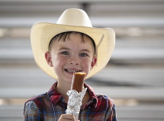 7-year-old Jackson Long enjoys a foot-long corn dog before taking part in Mutton Bustin’ at the  75th Annual Taylor Rodeo on Thursday, July 17,2025.  Coordinated by the Taylor Rodeo Association, the three-night rodeo is held at the Williamson County Expo Center.  Photo by Andy Sharp.  