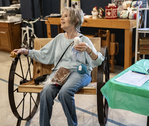 Sue Long relaxes in a custom-made wagon wheel chair created by  Ben Parker, a client auction item for sale at the 22nd Annual  Come Home for Christmas Celebration held at St. John’s Methodist Church on Saturday, November 22, 2025.    As of late Saturday morning, Sue was the highest bidder, hoping to make it part of her home.   The event included a Youth Cafe, with proceeds going to the St. John’s Youth Ministry.  There was also a silent auction, crafts made by church members, plus a bake sale.