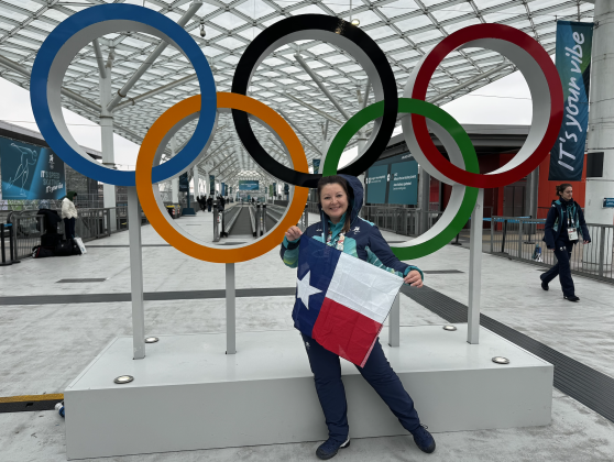 Leander resident Anna Yelaun stands in front of the Olympic rings holding a Texas flag. Ms. Yelaun volunteered at the 2026 Milan Cortina Winter Olympics as the lead volunteer for speed skating. (Photo courtesy Anna Yelaun)
