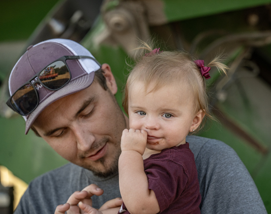 Montgomery Raesz, 26,  spends time with his daughter Maizlee, 16-months-old,  as they take a supper break before guiding  a combine through a field of corn along County Road 101 during the 2026 corn harvest of brothers Shaun and Steven Raesz on Saturday, July 11, 2026.  Montgomery is the first-born son of Shaun Raesz. The family is harvesting over 4,000 acres of corn this season.     Photo by Andy Sharp