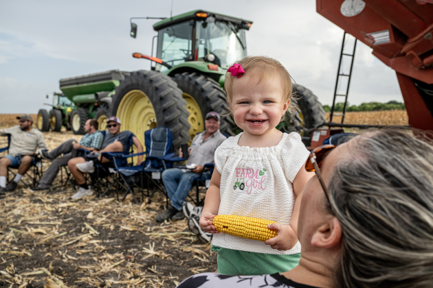 16-month-old Maizlee Raesz sits with her grandmother Kari Raesz as the farm crew takes a supper break  along County Road 414 during corn harvest of brothers Shaun and Steven Raesz  on Friday, July 10, 2026.  Kari is Shaun’s wife.  Maizlee is the daughter of Shaun’ and Kari’s  son Montgomery and his wife Madalynn.  The ear of corn she’s holding was just a play toy for Maizlee.   These East Williamson County farmers are harvesting over 4,000 acres of corn this year.    Photo by Andy Sharp