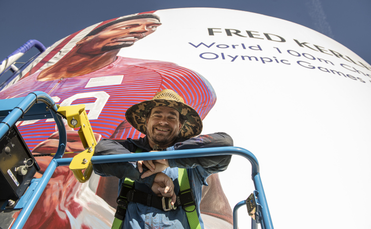 Adam Davenport poses for a photo on Thursday, January 19, with the mural he's painting of World 100 Meter champion and Olympics Silver medalist Fred Kerley, a Taylor native.   The mural is being painted on the Ford Ground Storage Tank at 1606 North Main Street, Taylor. Mr. Davenport says he has about ten days of work to complete the project.   The city will dedicate it during a ceremony on February 4 at 10 a.m. (Photos by Andy Sharp)