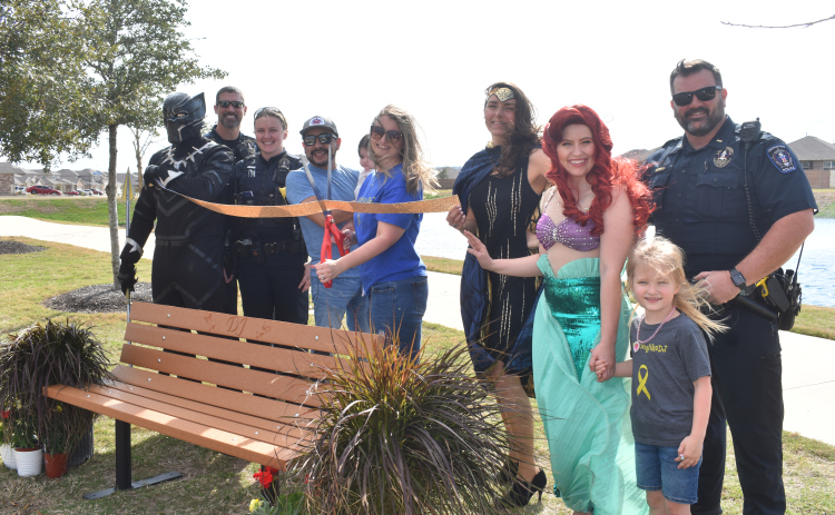 Amanda Torres, center, is surrounded by family and superhero friends as she cuts a ribbon to a bench dedicated to her late son, DJ Pruitt. Photo by Katherine Anthony